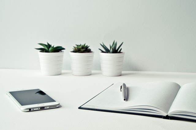 Three white ceramic pots with succulents in each one near an open notebook and a mobile phone